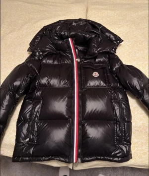 Moncler jacka  - Storlek: 5, passar både XL och L. Säljer en svart pufferjacka från Moncler med glansig finish och ikonisk logga på bröstfickan. Jackan har en fet röd och vit rand längs dragkedjan, hög krage och stora fickor. Perfekt för kalla dagar när du vill se både chill och lyxig ut. 