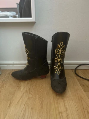 Annas Svarta Disney boots  - Unika svarta boots från Disney med spetsig tå och låg klack i vinrött. Skorna har snygga broderade detaljer i guld och paljetter på foten. Dragkedja på sidan för enkel påtagning. FÖR BARN! 