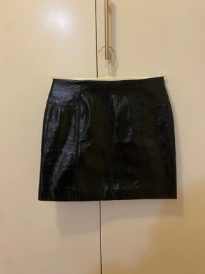 Svart minikjol i skinn - Säljer en svart, glansig minikjol i skinnimitation. Kjolen har en synlig dragkedja bak. Strl 34