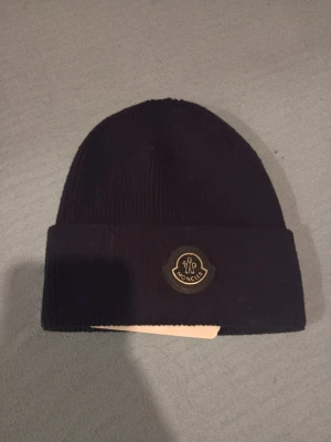 Svart Moncler mössa  - Svart ribbstickad mössa från Moncler med klassisk uppvikt kant och Moncler-logga framtill. Tillverkad i  bomull, perfekt för att hålla värmen och ge en clean look. Snygg och enkel att matcha med olika outfits.