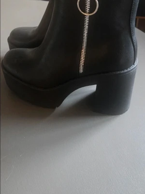 Svarta boots med klack och dragkedja - Säljer ett par svarta boots i storlek 39 med chunky klack och plattform. Skorna har rund tå och en snygg silverfärgad dragkedja med ringdetalj på sidan. Materialet är skinnimitation och sulan är rejäl för extra höjd och stabilitet.