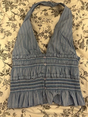 Blå topp - Toppen har vita ränder, volangdetaljer och knappar framtill. Smockad midja och halterneck gör den extra snygg. Perfekt för varma dagar och sommarhäng!