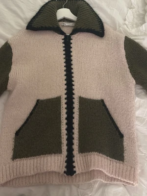 Stickad kofta från Zara i rosa och olivgrön - Säljer en stickad kofta från Zara i storlek S. Koftan har en ljusrosa bas med olivgröna ärmar och fickor, samt svarta detaljer längs dragkedjan och kragen. Modellen är långärmad med ribbade muddar och två stora fickor framtill. Perfekt för lager-på-lager under kyliga dagar.