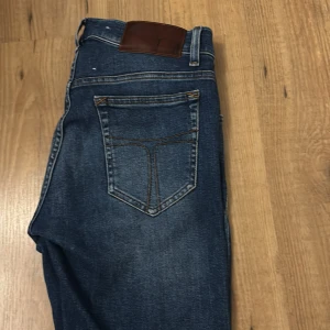 Tiger of Sweden blå jeans skinny fit - Snygga blå jeans från Tiger of Sweden med slim fit och klassisk femficksdesign. Jeansen har en mörkblå tvätt med lätt slitning och kontrastsömmar. Materialet är jeans med bomullskänsla och de har normal passform i midjan. Perfekta för dig som gillar stilrena och moderna jeans.