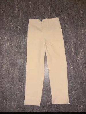 Beiga kostymbyxor från Zara - Snygga beiga kostymbyxor från Zara med hög midja och raka ben. Byxorna har diskreta sömmar framtill, dragkedja och knappstängning. Materialet känns mjukt och har en lätt stretch för extra komfort. Perfekta till en clean och stilren look.