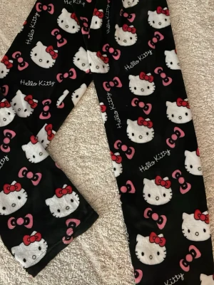 Hello Kitty mjukisbyxor svart/rosa - Supermysiga svarta mjukisbyxor med Hello Kitty-motiv och rosa rosetter över hela byxorna. Byxorna är i mjukt fleecematerial och har en loose passform, perfekta för chill dagar hemma.