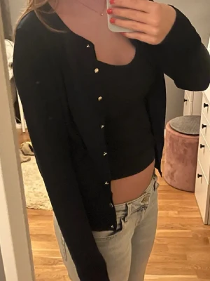 Svart kofta från Hollies & White - Snygg svart kofta från Hollies & White med rund hals och silvriga knappar framtill. Koftan har lång ärm och är tillverkad i mjuk bomull, perfekt att slänga över en topp eller linne. Enkel och stilren design som funkar till jeans eller kjol.
