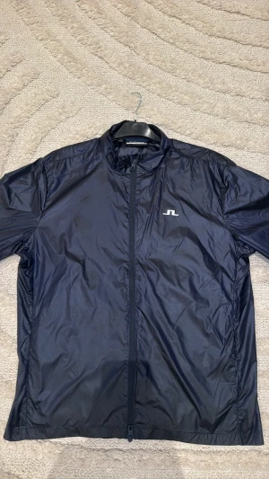 Mörkblå vindjacka från J.Lindeberg - Snygg mörkblå vindjacka från J.Lindeberg med stilren design och diskret logga på bröstet. Jackan har hel dragkedja framtill, ståkrage och är tillverkad i lätt polyester som är perfekt för blåsiga dagar. Passar dig som gillar sportig och clean look.