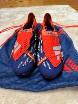 Adidas Predator blå/röda fotbollsskor - Säljer ett par Adidas Predator fotbollsskor i storlek 43 1/3, skruvdobbar, använda 3-4 gånger nypris 3199. Kommer med matchande blå/röd påse.  