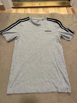Grå Adidas t-shirt med svarta ränder - Snygg grå t-shirt från Adidas i storlek XS med klassiska svarta ränder längs axlarna och ett svart Adidas-tryck på bröstet. T-shirten har rund hals och korta ärmar, perfekt för en sportig och chill stil. Materialet är mjuk bomull som känns skönt mot huden.