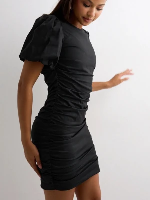 Svart kort klänning med puffärm - Säljer en svart kort klänning med puffiga korta ärmar och rynkade sidor. Klänningen har en figurnära passform och är sydd i ett slätt tyg som ger en snygg siluett. Perfekt för dig som vill sticka ut med en trendig look. Från Nelly med öppen rygg och dragkedja baktill