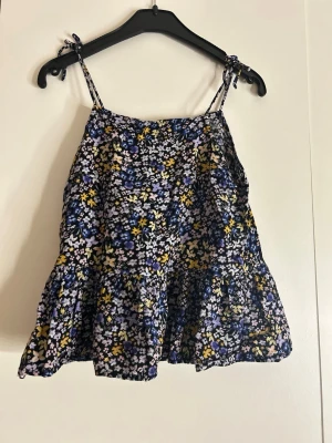 Blommig linnetopp från H&M storlek 140 - Supersöt linnetopp från H&M storlek 140 med blommigt mönster i svart, lila, blått och gult. Toppen har tunna axelband med knyt och en luftig, volangig nederdel. Perfekt för varma dagar och ger en riktigt somrig vibe.