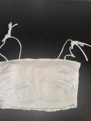 Vit bandeau topp från Zara - Säljer en vit bandeau topp från Zara i storlek S. Toppen har smala axelband med knyt och en smockad baksida med volangkant. Materialet är mjukt och har en lätt glansig yta som ger en fräsch känsla. Perfekt för varma dagar och enkel att matcha.