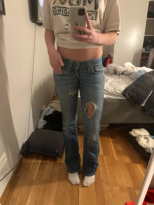 Replay bootcut jeans med slitningar - Säljer ett par blå Replay jeans med bootcut-snitt och låg midja. Jeansen har slitna detaljer och hål på benen för en cool, avslappnad look. Klassisk femficksmodell med dragkedja och knapp. Perfekta för dig som gillar streetstyle.