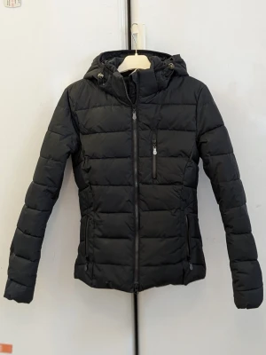 Ridjacka,  JH Collection  - Ridjackan har använts en enda gång. Snygg svart ridjacka, vinterjacka, pufferjacka med huva och dragkedja framtill. Jackan har quiltad design, två sidofickor med dragkedja och en bröstficka med dragkedja. Perfekt ridjacka för kyliga dagar och enkel att matcha med olika outfits.