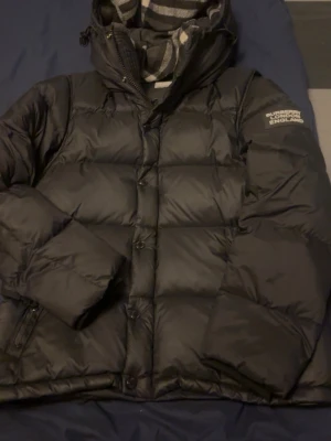 Svart pufferjacka från Burberry - Svart pufferjacka från Burberry London England med rutigt foder i huvan. Jackan har både dragkedja och knappar framtill, samt långärmade ärmar. Perfekt för kalla dagar med sin tjocka och quiltade design.