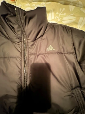 Svart pufferjacka från Adidas - Svart pufferjacka från Adidas med hög krage och synlig dragkedja framtill. Jackan har Adidas-loggan på bröstet och är tillverkad i polyester. Perfekt för kalla dagar när du vill hålla dig varm och samtidigt se sportig ut.