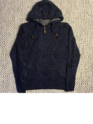Mörkblå stickad hoodie med dragkedja - Säljer en mörkblå stickad hoodie med snyggt kabelmönster och huva. Tröjan har dragkedja framtill, snörning vid huvan och ribbade muddar vid ärmslut och nederkant. Perfekt för kyliga dagar och riktigt mysig att ha på sig.