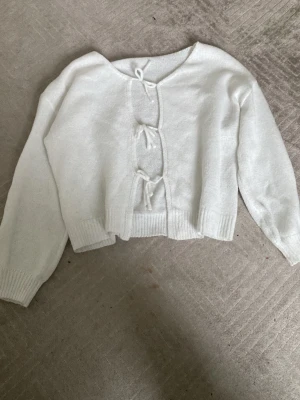 Vit stickad kofta med knyt - Mysig vit stickad kofta med långa ärmar och tre knyt framtill istället för knappar. Koftan har ribbade muddar vid ärmslut och nederkant, samt en något croppad passform. Perfekt för lager-på-lager och chill stil.