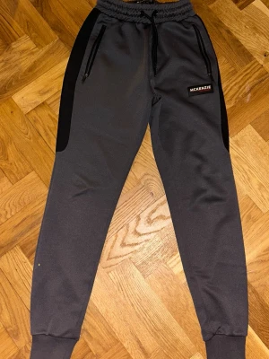 Mörkgrå mjukisbyxor från McKenzie - Säljer ett par mörkgrå mjukisbyxor från McKenzie med svarta sidopaneler, snörning i midjan och dragkedjeförsedda fickor. Byxorna har ribbade muddar vid bensluten och en stilren logga på ena benet. Perfekta för chill eller träning.