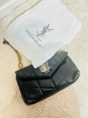 Svart quiltad axelväska från YSL - Säljer en svart quiltad axelväska från Yves Saint Laurent med guldfärgade kedjedetaljer och YSL-logga framtill. Väskan är tillverkad i skinn och har ett innerfack med dragkedja. Perfekt storlek för det viktigaste och riktigt snygg design.