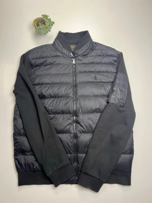 Svart Cardigan/hybrid jacka från Polo Ralph Lauren - Svart cardigan  från Polo Ralph Lauren med quiltad framsida och rygg, samt släta ärmar i bomull. Jackan har dragkedja framtill och en cool ficka med dragkedja på ena ärmen. Insidan har ett rutigt foder. Perfekt för dig som gillar stilren och klassisk look.