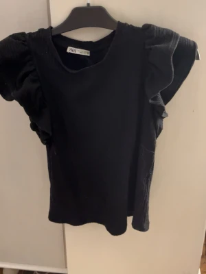 Svart ribbad topp med volangärmar från Zara - Säljer en svart ribbad topp från Zara i storlek S. Toppen har korta volangärmar som ger en cool och trendig look. Materialet är mjukt och stretchigt, perfekt för att styla med jeans eller kjol. Enkel rund halsringning och snygg passform.