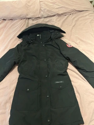 Svart parkajacka från Canada Goose - Svart parkajacka från Canada Goose med ikonisk röd och blå patch på ärmen. Jackan har huva med snörning, stora fickor framtill och knappar längs hela fronten. Materialet är slitstark polyester och dunfyllning för extra värme. Perfekt för kalla vinterdagar. päls ingår med jackan är oanvänd 