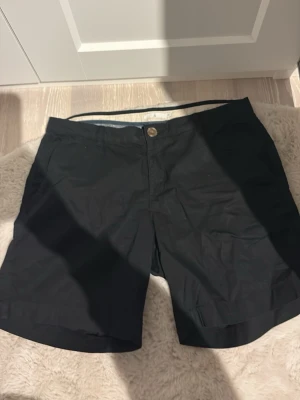 Marinblåa shorts med knapp - Säljer ett par marinblåa shorts från Åhléns som knappt är använda. Det finns en dragkedja med en knapp vid mitten av shortsen. 