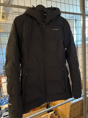 Svart dunjacka från 67°N åhkká - Svart dunjacka från 67°N åhkká med huva och dragkedja framtill. Jackan har två sidofickor med dragkedja och ett snyggt mönstrat innerfoder. Perfekt för kalla dagar och har en clean, stilren look.