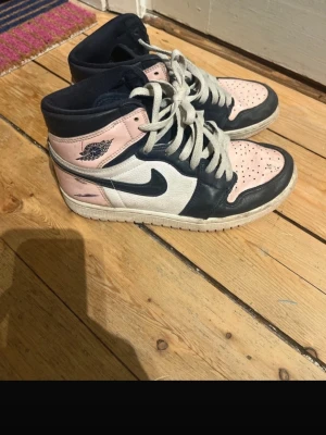 Nike Jordan 1 High i rosa och marinblå - Nike Jordan 1 High sneakers med snygg färgkombo i ljusrosa, marinblå och vit. Klassisk snörning och rund tå, tillverkade i skinn med vit platt sula. Ikonisk vinge-logga på sidan och svart swoosh. Perfekt för dig som gillar streetwear och vill sticka ut.
