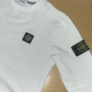 Vit Stone Island sweatshirt - Vit långärmad sweatshirt från Stone Island med klassisk rund hals och ikoniska logopatchar på bröstet och ärmen. Tröjan är i mjuk bomull och har en clean, stilren look som passar perfekt till streetwear-stilen.