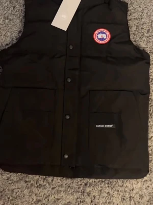 Svart och grå dunväst från Canada Goose - Två stilrena dunvästar från Canada Goose, en i svart och en i grått. Båda har hög krage, tryckknappar framtill, två stora fickor och klassisk Canada Goose-logga på bröstet. Materialet är slitstark polyester och dunfyllning som håller dig varm. Perfekt för kalla dagar och snygg till streetwear.