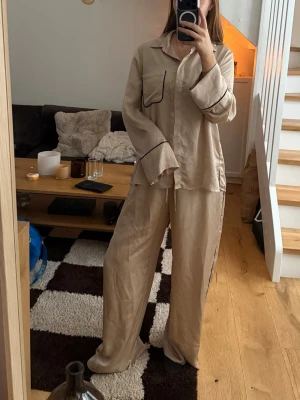 Beige pyjamasset från H&M Home - Säljer ett beige pyjamasset från H&M Home i storlek S. Setet består av en skjortliknande överdel med svarta kantdetaljer och en bröstficka, samt matchande vida byxor med dragsko i midjan. Materialet är mjukt och glansigt polyester, perfekt för chillkvällar.