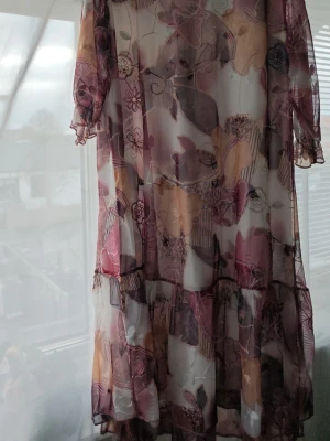 Blommig klänning str.52  - Säljer en romantisk långklänning från Pelageia med transparent tyg och blommigt mönster i lila, beige och rosa toner. Klänningen har volangdetaljer och trekvartsärmar med lätt rynkning. Materialet är 65% bomull och 35% spandex för en mjuk och följsam känsla.