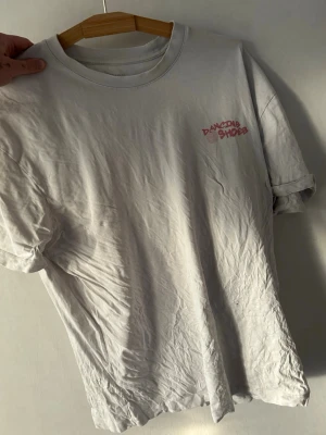 Vit Dancing Shoes t-shirt - Vit t-shirt från Dancing Shoes med tryck i rött fram och bak. Framsidan har en liten logga på bröstet och baksidan har ett stort grafiskt tryck med smileys och text. Klassisk passform och rund hals. Perfekt för dig som gillar streetwear vibes.