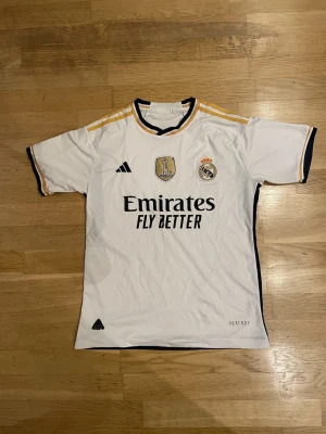Real Madrid matchtröja 2024 Adidas - Säljer en officiell Real Madrid matchtröja från säsongen 2024 med FIFA World Champions-märke. Tröjan är vit med guld- och marinblå detaljer, har klubbmärke och Adidas-logga på bröstet samt 'Emirates Fly Better' tryck. Perfekt för dig som älskar fotboll och Real Madrid.