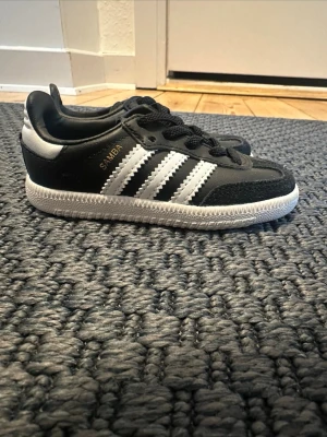 Adidas Samba sneakers svart/vit st 21 - Säljer ett par klassiska Adidas Samba sneakers i svart och vitt med de ikoniska tre ränderna på sidan och blå Adidas-logga på plösen. Skorna har snörning, rund tå och platt sula. Materialet är en mix av skinn och syntet, perfekt för dig som gillar retrostil.
