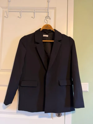 Svart kavaj från Mango - Stilren svart kavaj från Mango med klassisk krage och två fickor framtill. Kavajen har lång ärm och är gjord i ett mjukt polyestermaterial som sitter snyggt och bekvämt.