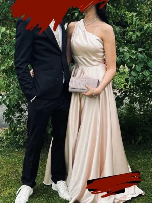 Champagnefärgad oneshoulder långklänning - Säljer en elegant långklänning i champagnefärg med oneshoulder-design och markerad midja. Klänningen har ett vackert fall och är tillverkad i ett glansigt material som ger en lyxig känsla. Perfekt för bal eller festliga tillfällen.