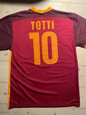 AS Roma Totti 10 matchtröja - Säljer en snygg AS Roma matchtröja med Totti och nummer 10 på ryggen. Tröjan är vinröd med mörkröda och orange detaljer, samt klubbmärke på bröstet. Materialet är polyester och har en klassisk v-ringad krage. Perfekt för dig som älskar fotboll och italiensk stil. Lite bruksslitage i armhålorna men annars bra skick