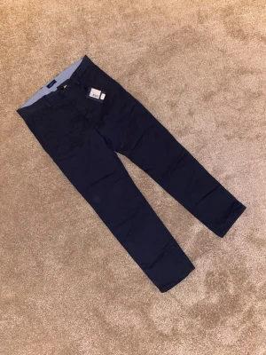 Mörkblå chinos från GANT - Snygga mörkblå chinos från GANT med klassisk passform och stilrena detaljer. Byxorna har bälteshällor, framfickor och är tillverkade i mjuk bomull. Perfekta för dig som gillar en clean och enkel look. Lappen sitter kvar och storleken är W32 L32.