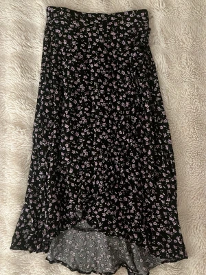 Svart blommig långkjol från Lindex - Svart långkjol från Lindex med små lila och rosa blommor över hela tyget. Kjolen har en mjuk och flowy form med lätt volang nedtill. Perfekt för dig som gillar romantiska och söta detaljer. Resår i midjan för skön passform.
