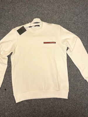 Vit sweatshirt från Prada - Säljer en stilren vit sweatshirt från Prada med rund hals och lång ärm. Tröjan har en diskret logga i rött och svart på bröstet. Mjuk och skön bomullskvalitet, perfekt för dig som gillar minimalistisk design och exklusiva märken.