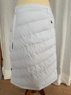 Oanvänd vit quiltad kjol från Dobsom  - Quiltad vit kjol från Dobsom med dragkedja i sidan och bälteshällor. Kjolen är tillverkad i 100% polyester och har en slits bak för extra rörelsefrihet. Perfekt för kalla dagar när du vill hålla dig varm och ändå se fräsch ut. Ca 65 cm lång, se bilder