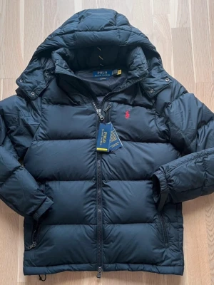 Svart pufferjacka från Polo Ralph Lauren - Svart pufferjacka från Polo Ralph Lauren med huva och röd logga på bröstet. Köptes för 4595kr ifrån ralph lauren butik. Materialet är polyester och dun, perfekt för kalla dagar. Snygg och stilren look. Kvitto finns!