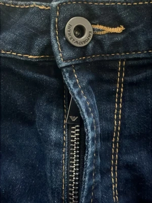 Emporio Armani mörkblå jeans, strl 32 - Snygga mörkblå jeans från Emporio Armani med klassisk rak passform och kontrastsömmar. Byxorna har dragkedja, knapp med logga och metallmärke på bakfickan. Tillverkade i jeansmaterial, passar dig som gillar stilrena detaljer och exklusiv känsla.