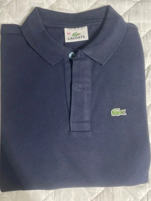 Mörkblå Lacoste pikétröja - Säljer en mörkblå pikétröja från Lacoste med klassisk krage och två knappar framtill. Tröjan har korta ärmar och är gjord i bomull. Den ikoniska gröna krokodilen är broderad på bröstet. Perfekt för dig som gillar stilrena och sportiga plagg.
