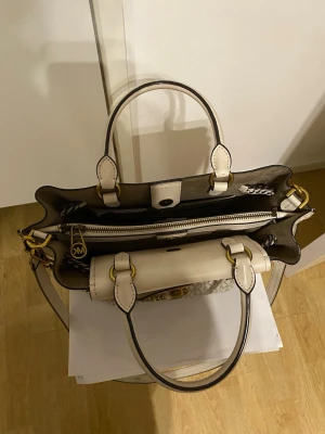 Beige MK handväska med kedja - Säljer en snygg handväska från Michael Kors i beige och vitt med MK-logga över hela väskan. Väskan har guldfärgade detaljer, kedja, flätad dekor och ett hänglås med MK-emblem. Handtag och axelrem i skinn, dragkedja upptill och flera fack inuti.
