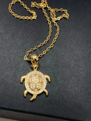 Guldig halsband med sköldpaddshänge - Snyggt halsband i guldton med ett hänge format som en sköldpadda, täckt av glittrande stenar. Kedjan är tunn och elegant, och hänget har fina detaljer med små stenar som ger extra bling. Perfekt accessoar för att lyfta din outfit.